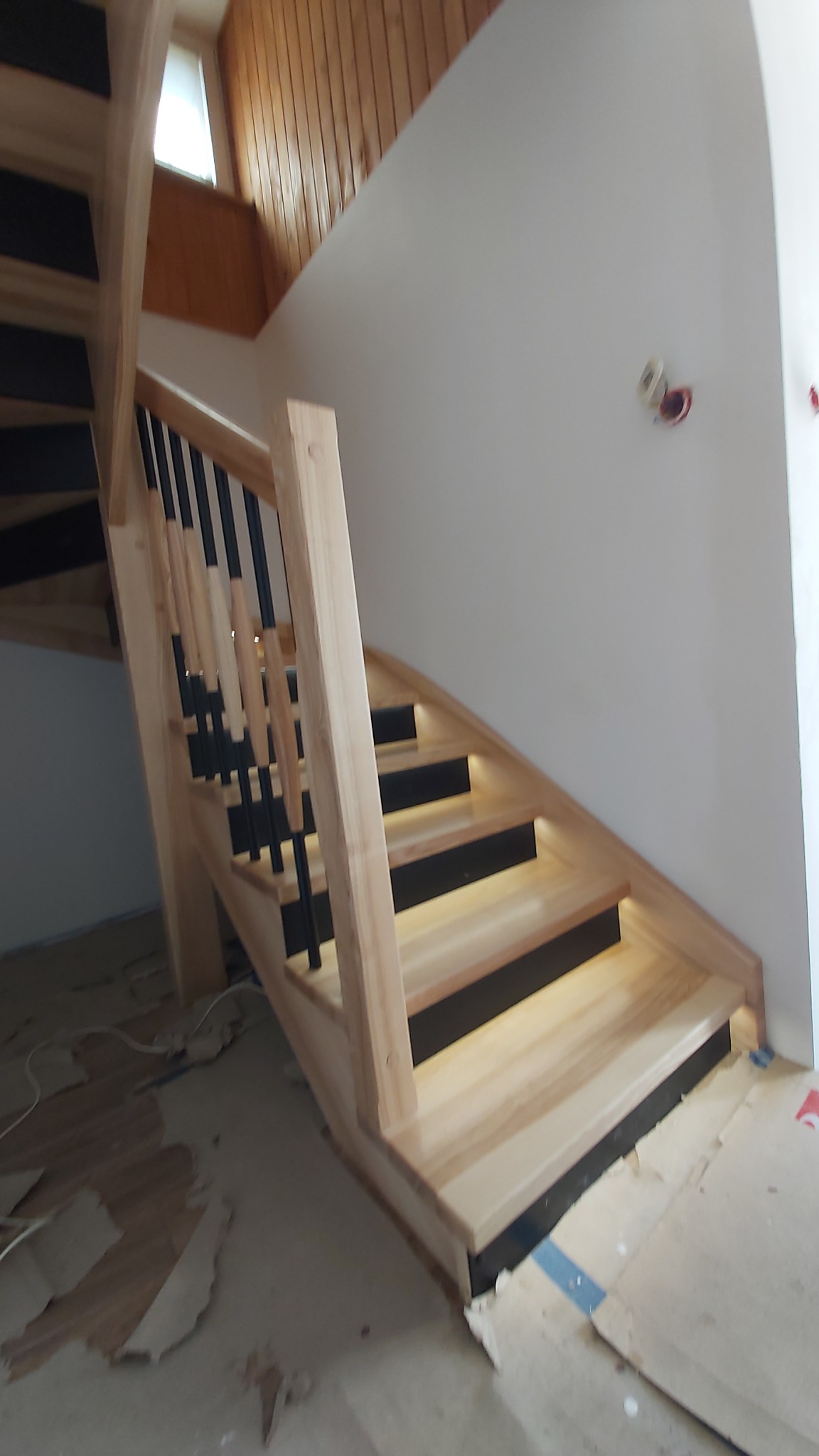 Jasne, drewniane schody z czarnymi akcentami i nowoczesną balustradą. Widoczne niedokończone wykończenie podłogi. Ujęcie z dołu, podkreślające detale konstrukcji.