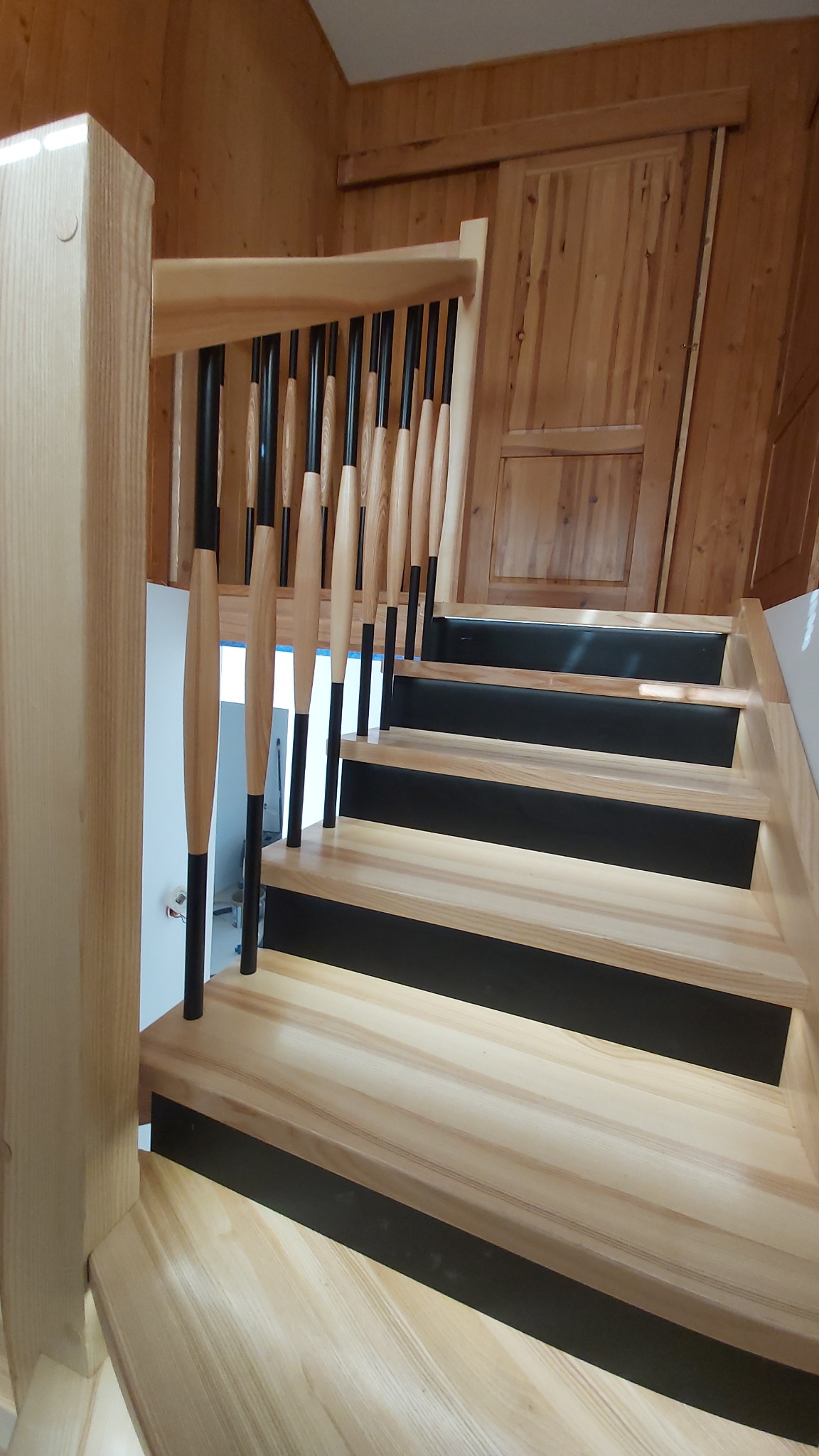Jasne, drewniane schody z czarnymi podstopnicami i balustradą z czarno-drewnianymi szczeblami, widok z dołu. Ściana wykończona drewnianą boazerią.
