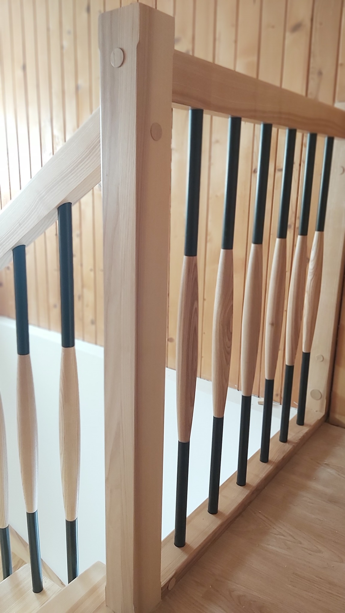 Jasne drewno i czarne elementy nowoczesnej balustrady schodowej z ozdobnymi wrzecionami, widok z dołu, drewniana boazeria w tle.