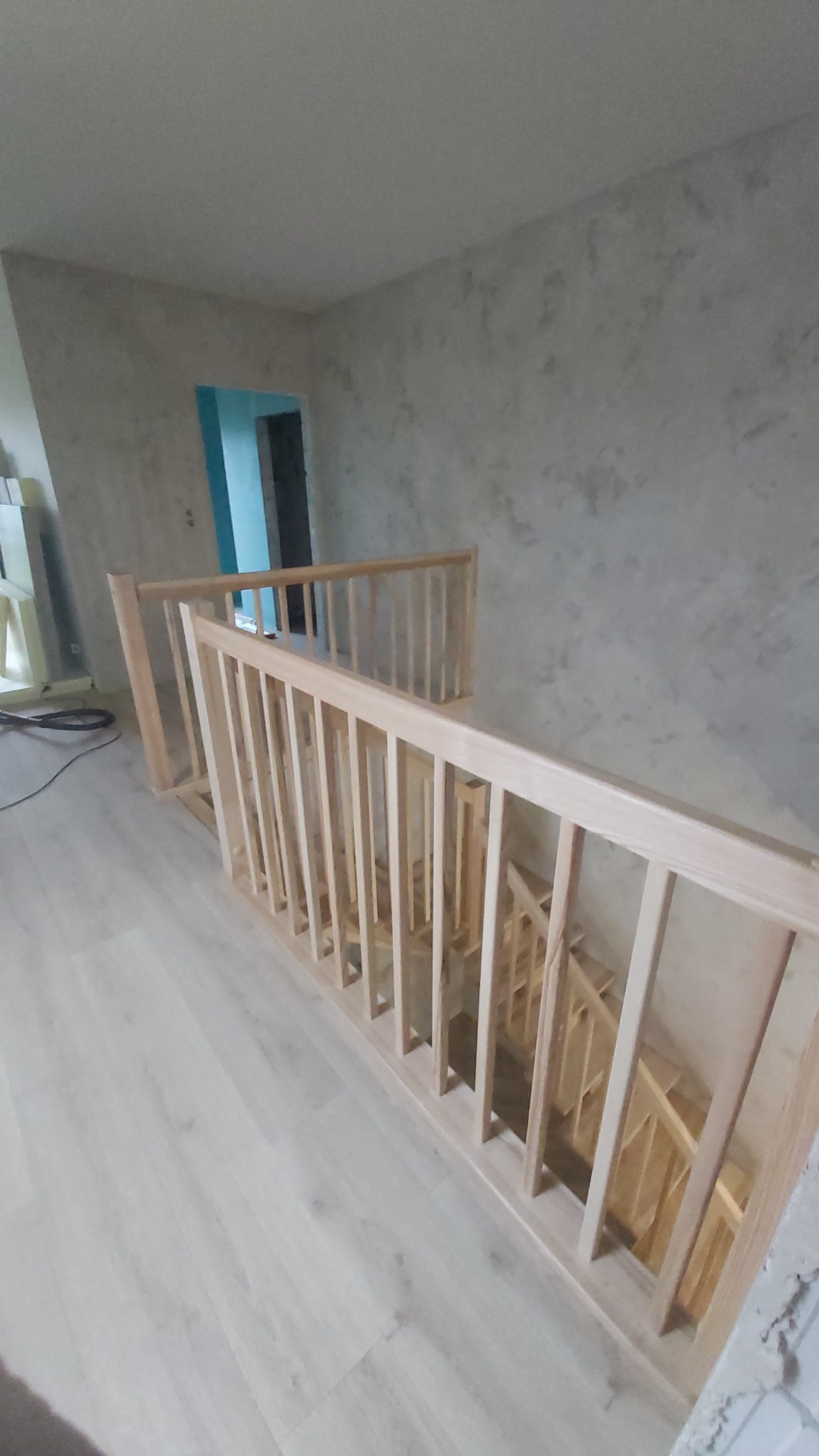 Jasne, surowe drewno balustrady schodowej w trakcie montażu, widok z góry. W tle ściany w odcieniach szarości i błękitne drzwi. Podłoga w jasnym odcieniu drewna.