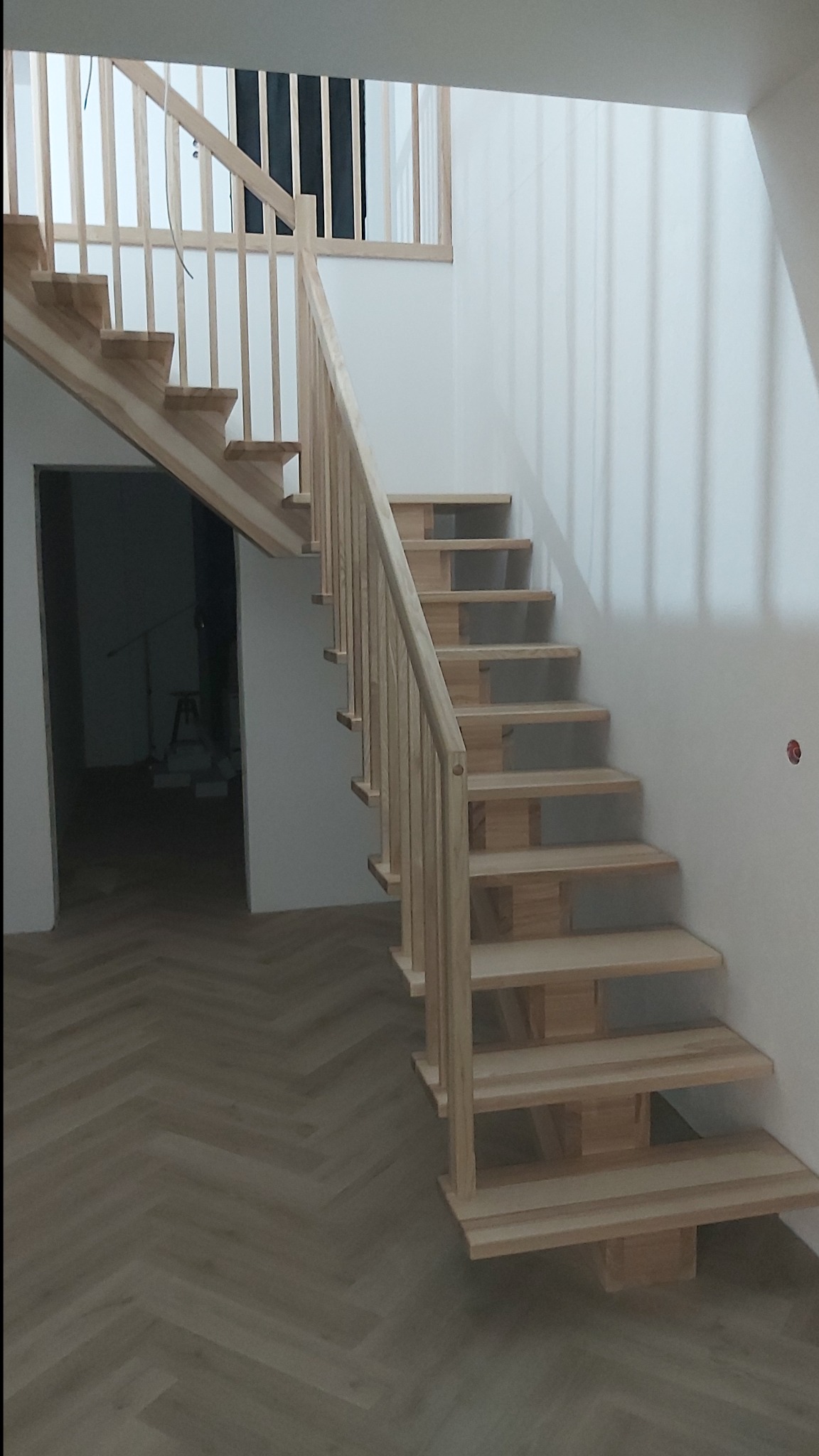 Jasne, drewniane schody z balustradą w nowoczesnym domu. Widoczny parkiet ułożony w jodełkę i fragment antresoli. Naturalne światło podkreśla prostotę i elegancję.