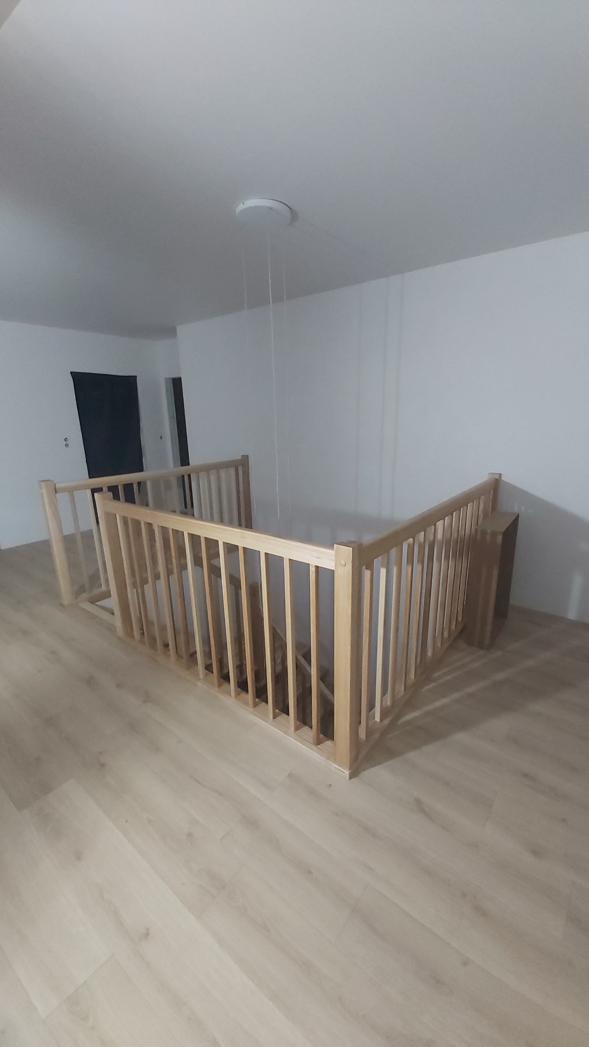 Jasna, drewniana balustrada schodowa z pionowymi szczebelkami, widoczna z góry, na tle białych ścian i podłogi z paneli. Minimalistyczny design, naturalne światło.