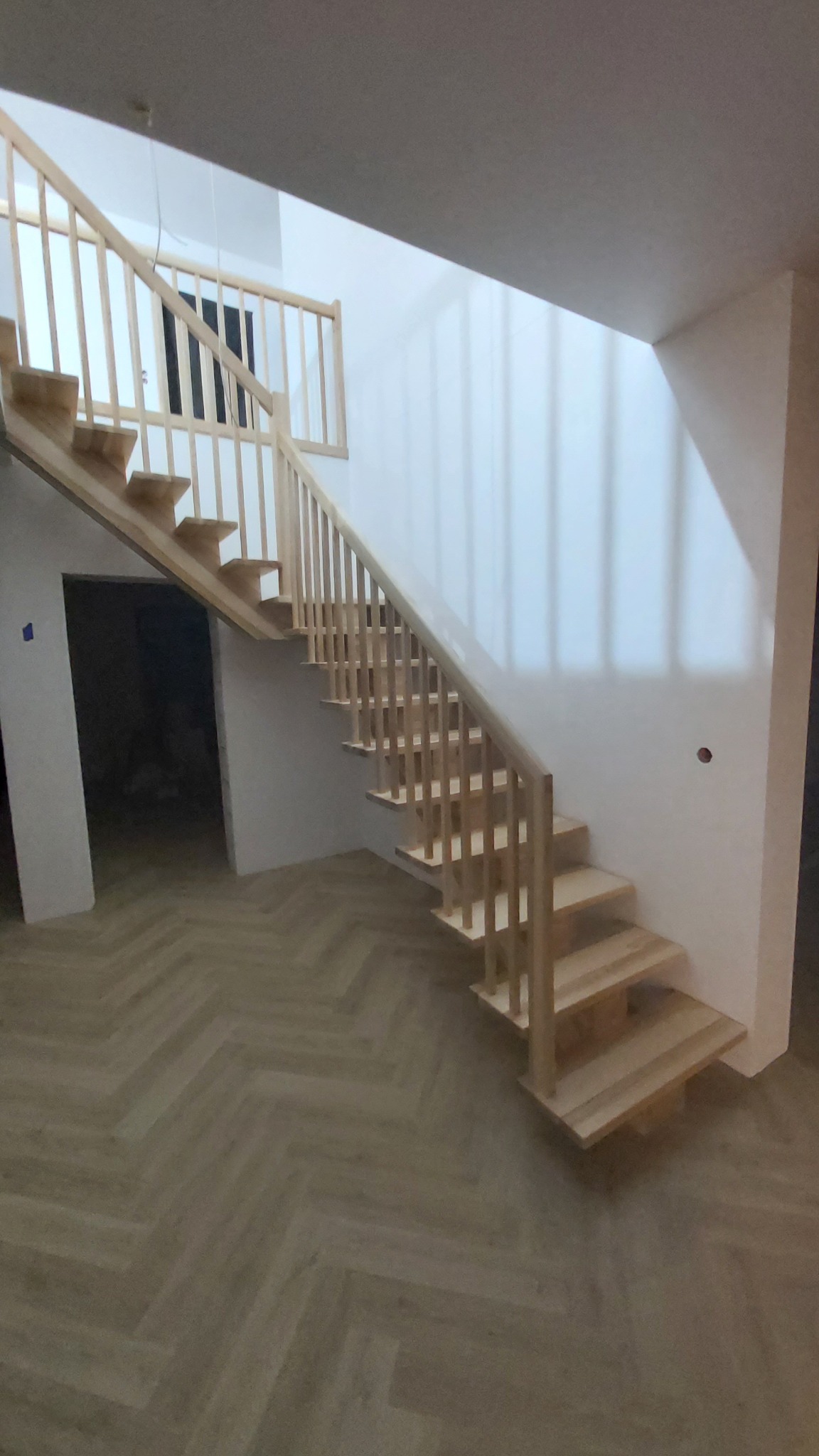 Jasne schody z balustradą wewnątrz budynku, z widocznym parkietem ułożonym w jodełkę. Naturalne światło podkreśla surowy styl wykończenia.