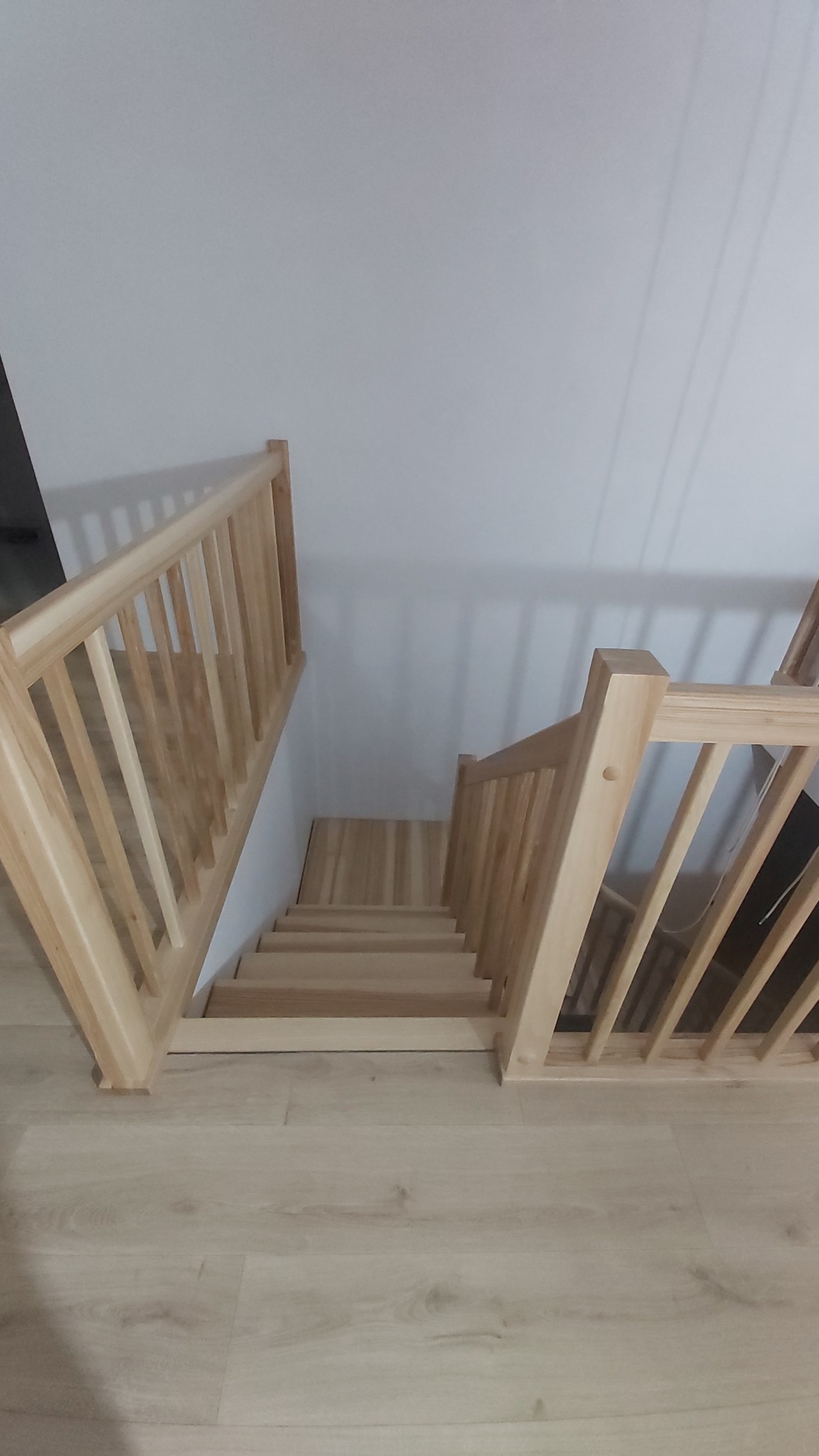 Jasne, drewniane schody z balustradą, widziane z góry. Naturalne oświetlenie podkreśla strukturę drewna. Elementy minimalistyczne, proste stopnie.