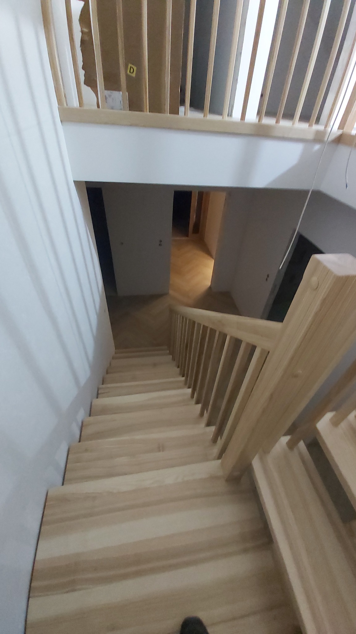 Jasne, drewniane schody wewnętrzne z balustradą, widok z góry. W tle parkiet ułożony w jodełkę i białe ściany. Elementy wykończeniowe w surowym drewnie.