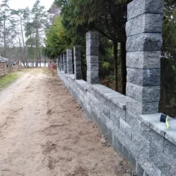 Nowo wybudowany, długi, szary mur z blok&oacute;w betonowych z regularnymi słupkami, biegnący wzdłuż piaszczystej drogi w kierunku jeziora, z widocznymi drzewami w tle i szczotką do czyszczenia na murze.