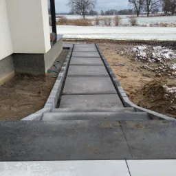 Ułożona ścieżka z betonowych płyt i kostki brukowej prowadząca do domu, widoczny teren przygotowany pod nasadzenia, w tle pole z resztkami śniegu i drzewa.