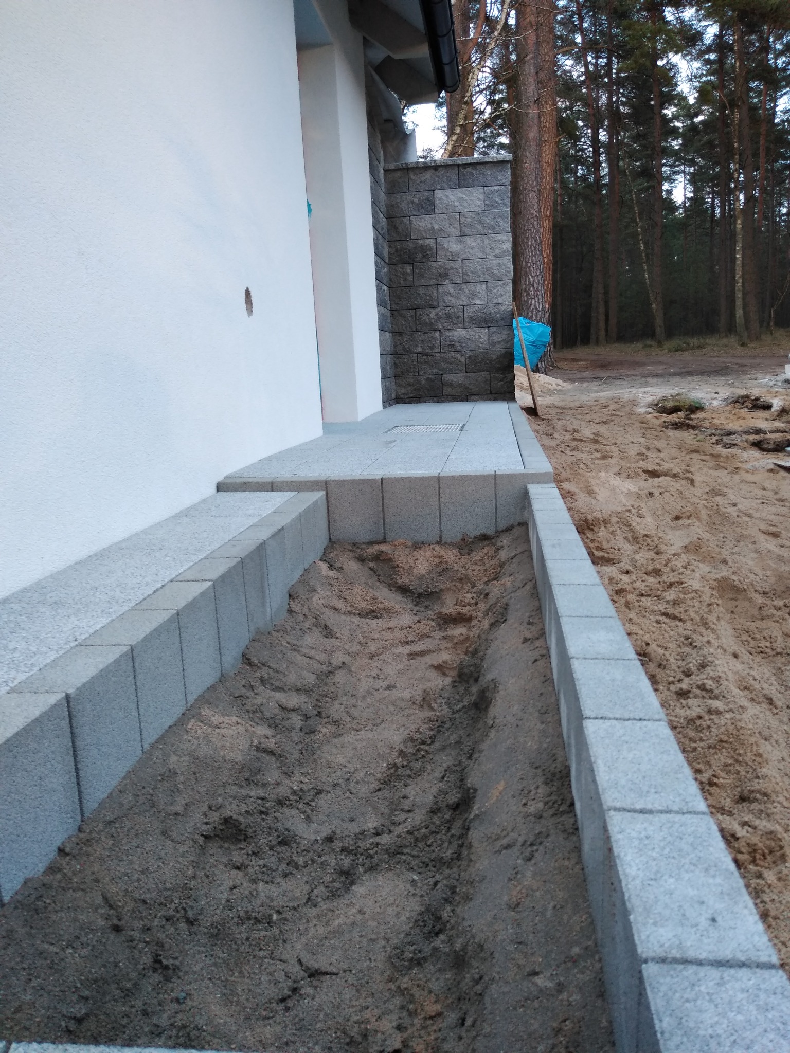 Przygotowanie podłoża pod kostkę brukową z obrzeżami, widoczna warstwa piasku i ziemi, w tle ściana budynku i las.
