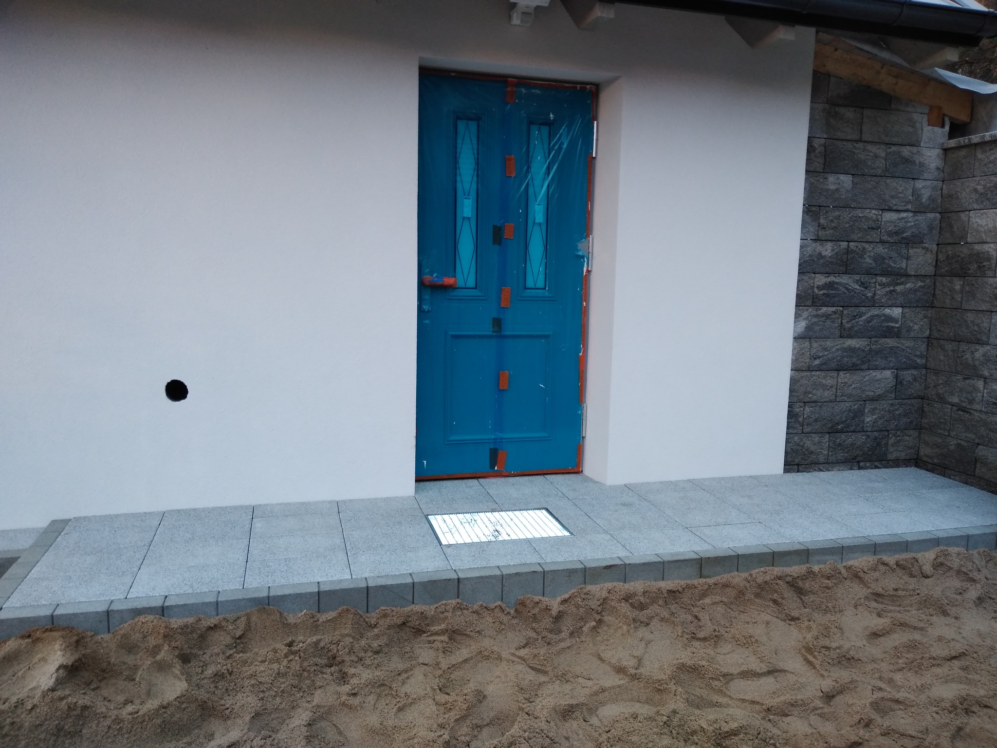 Świeżo ułożona, szara kostka brukowa przed białym budynkiem z niebieskimi drzwiami zabezpieczonymi folią, widoczna studzienka kanalizacyjna i pryzma piasku.