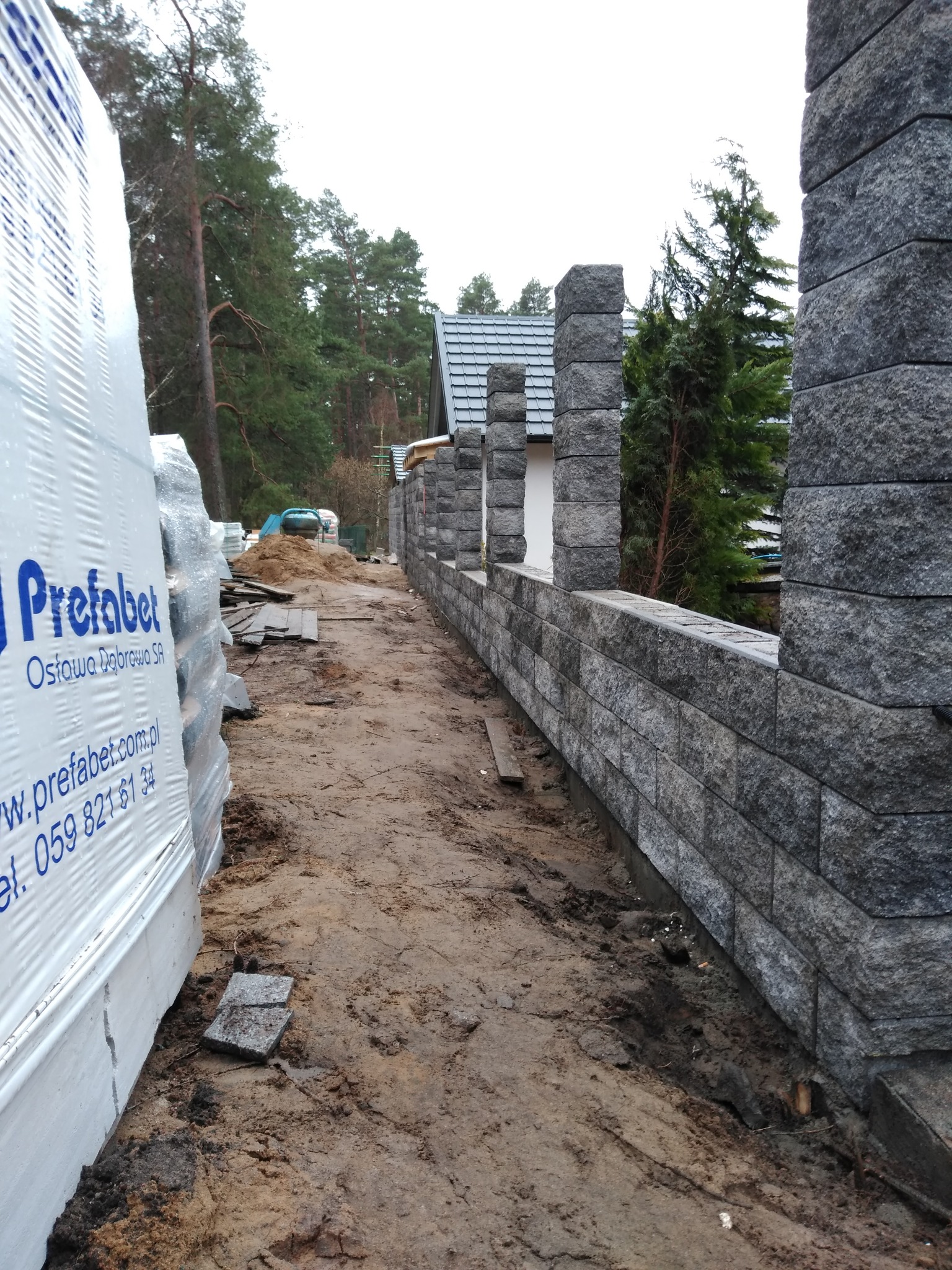Wznoszenie ogrodzenia z szarej, łupanej kostki granitowej; widoczny materiał budowlany w opakowaniu z logo producenta, ziemiste podłoże i fragment posesji w tle.
