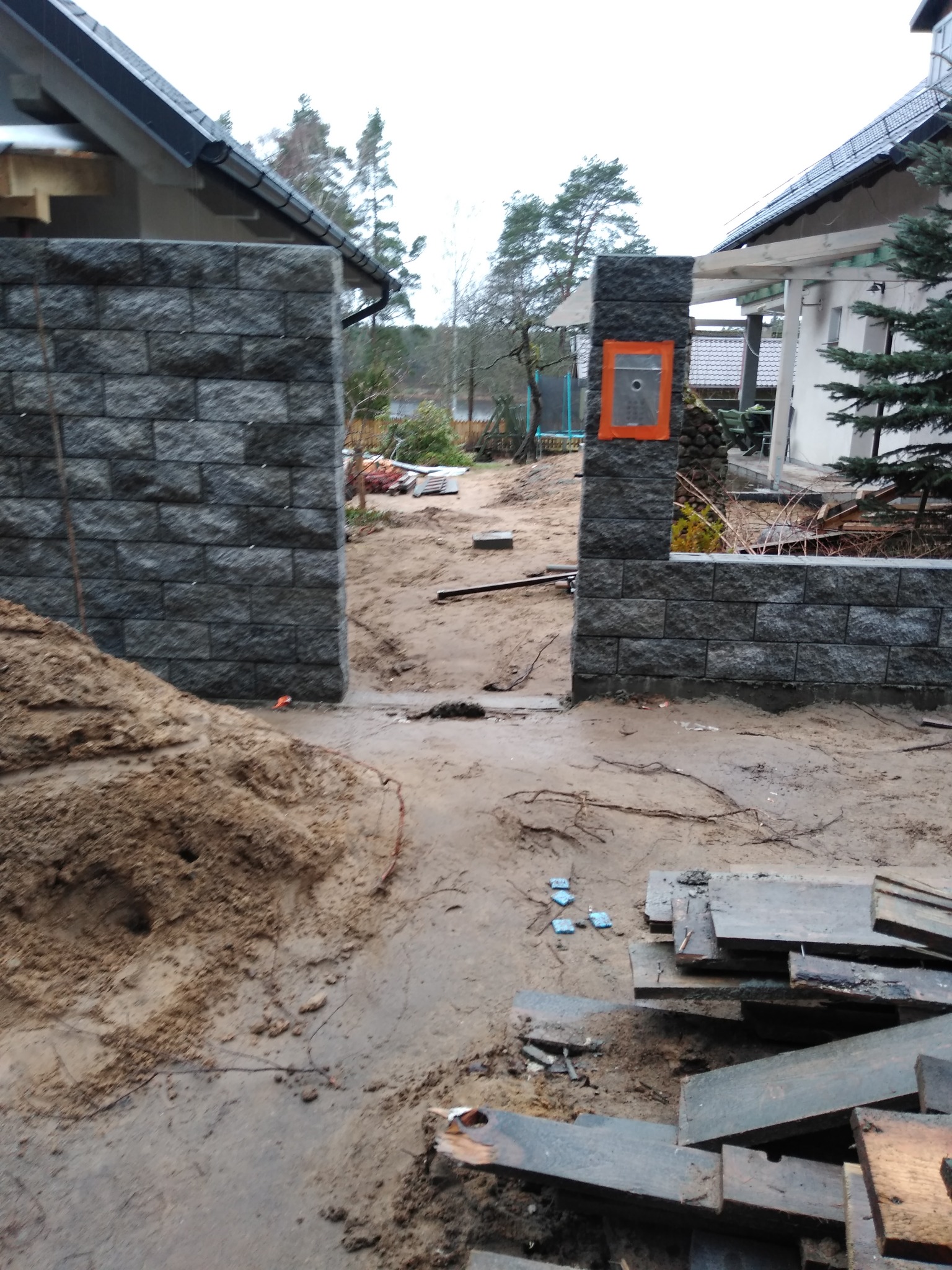 Fragment ogrodzenia z szarej kostki, z widocznym otworem na domofon zabezpieczonym pomarańczową taśmą, w trakcie budowy, z ziemią, deskami i elementami konstrukcyjnymi w tle.