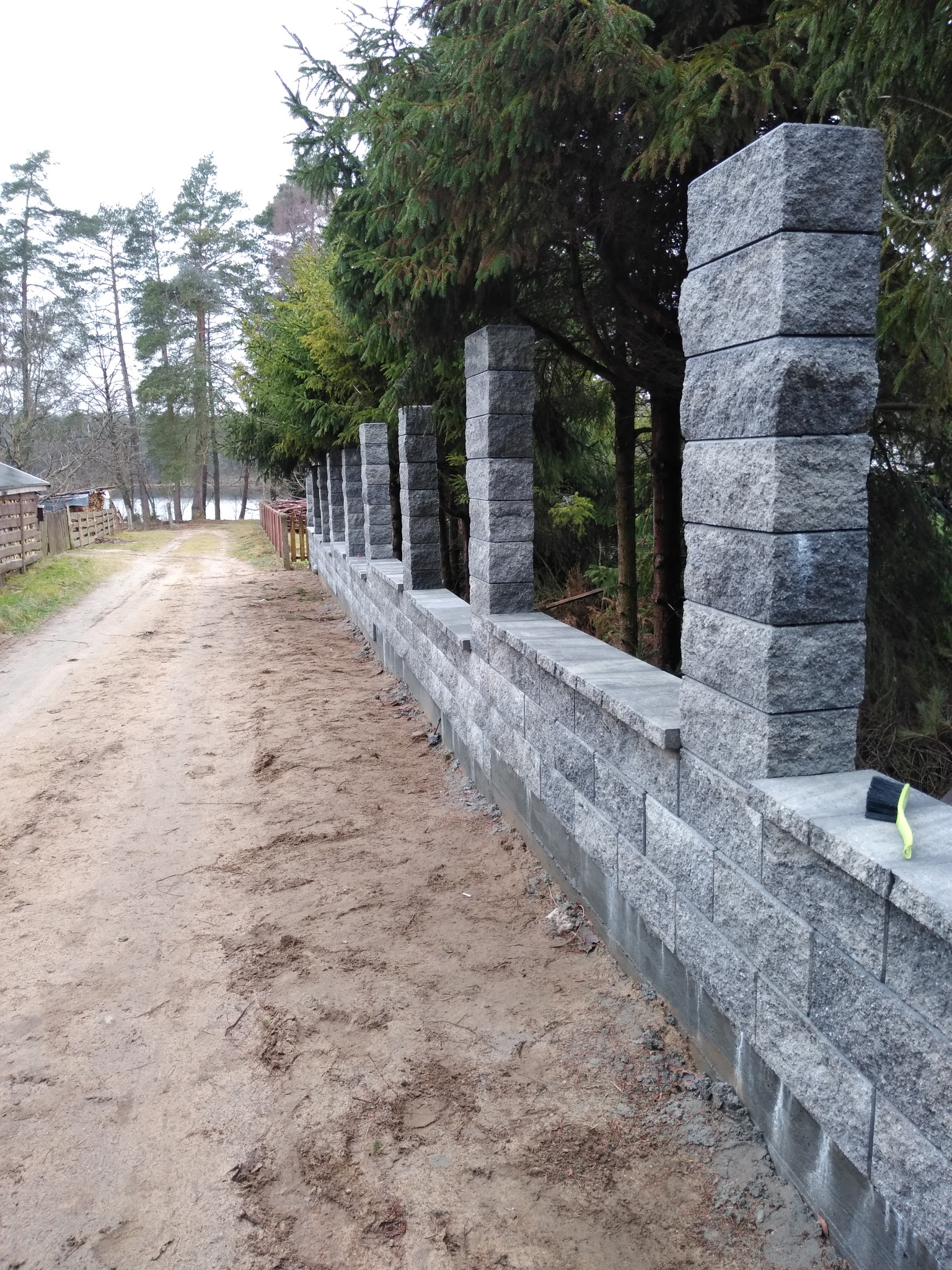 Nowo wybudowany, długi, szary mur z bloków betonowych z regularnymi słupkami, biegnący wzdłuż piaszczystej drogi w kierunku jeziora, z widocznymi drzewami w tle i szczotką do czyszczenia na murze.
