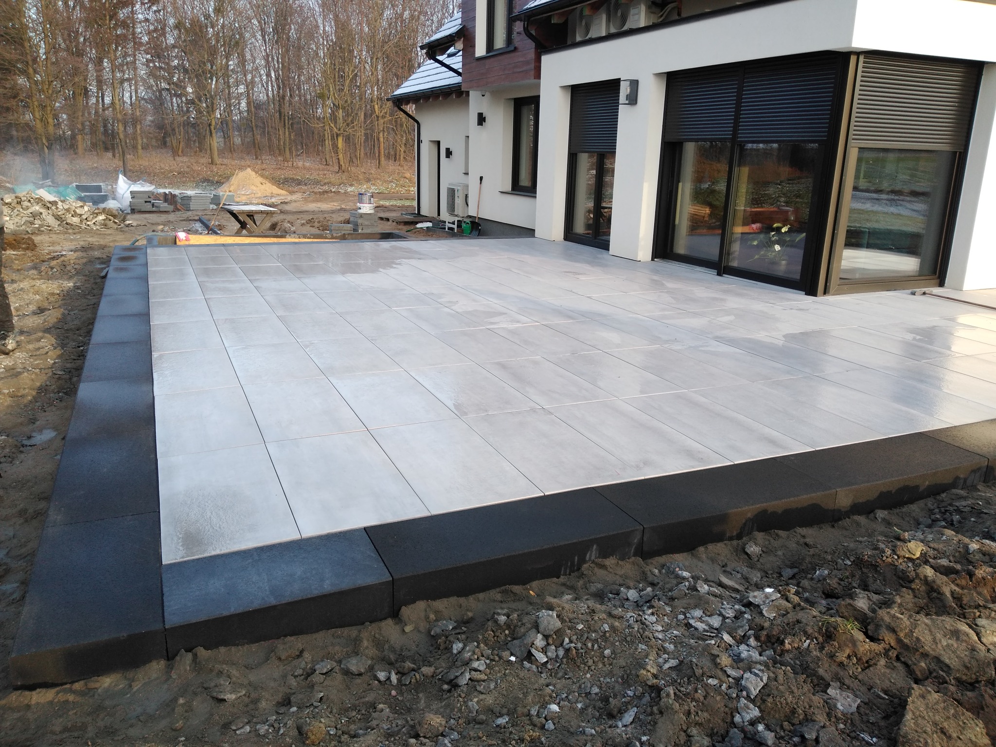 Nowoczesny taras z jasnych, kwadratowych płyt na zewnątrz domu, obrzeża z ciemnych kostek, widoczny teren przed domem w trakcie prac ziemnych.