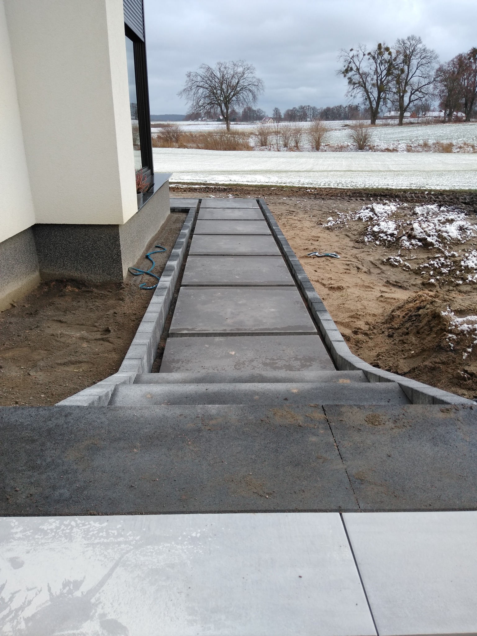 Ułożona ścieżka z betonowych płyt i kostki brukowej prowadząca do domu, widoczny teren przygotowany pod nasadzenia, w tle pole z resztkami śniegu i drzewa.