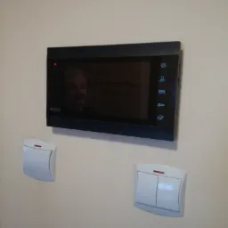 Zamontowany wideofon Eura na ścianie obok dwóch włączników światła; widoczny odbity obraz osoby w ekranie urządzenia.