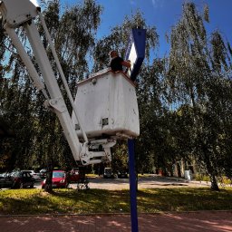 Tomek - Pracownik w koszu podnośnika teleskopowego, naprawiający lub czyszczący tablicę informacyjną na tle drzew i błękitnego nieba.