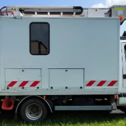 Biały samochód z podnośnikiem koszowym na tle budynków z panelami słonecznymi, widok z boku na trawniku.
