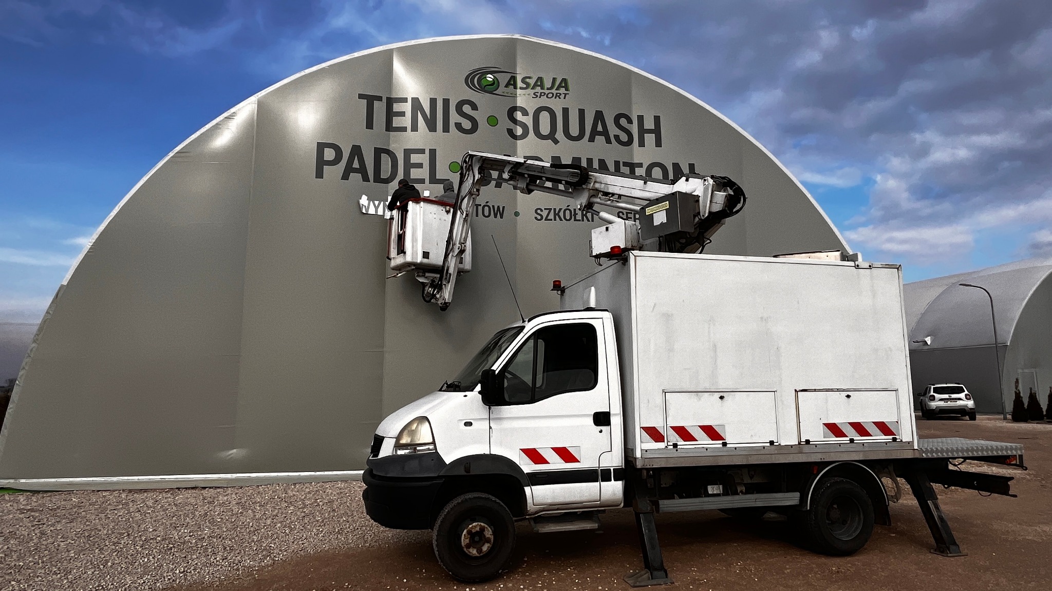 Biały samochód z wysięgnikiem podnośnikowym przy szarym namiocie sportowym z napisem 'Tenis Squash Padel Badminton', dwóch pracowników w koszu na wysięgniku, prace na wysokości