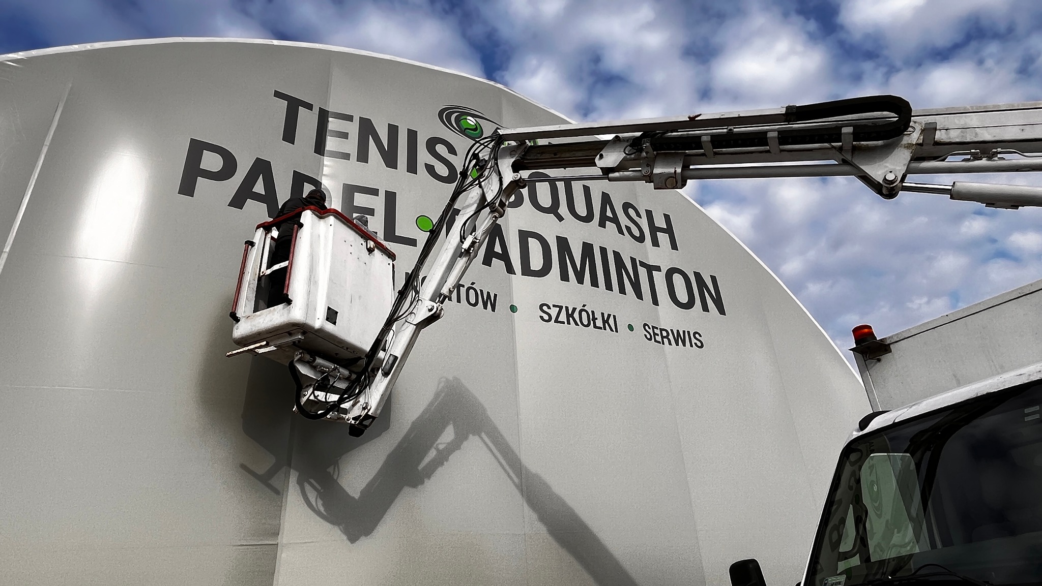 Podnośnik koszowy z pracownikiem na wysokości podczas montażu lub czyszczenia dużego baneru reklamowego z napisem: Tenis, Padel, Squash, Badminton, szkoły i serwis