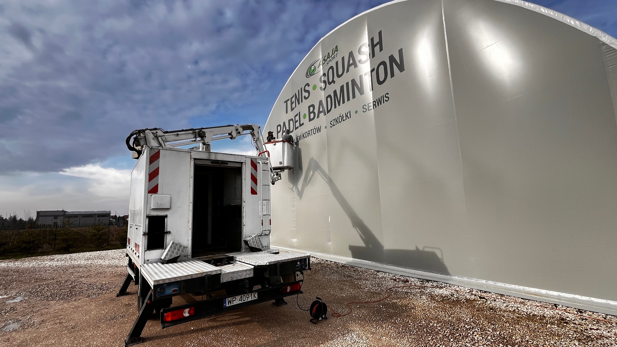 Pracownik w koszu podnośnika montuje lub czyści duży, biały namiot sportowy z napisem 'TENIS SQUASH PADEL BADMINTON'.