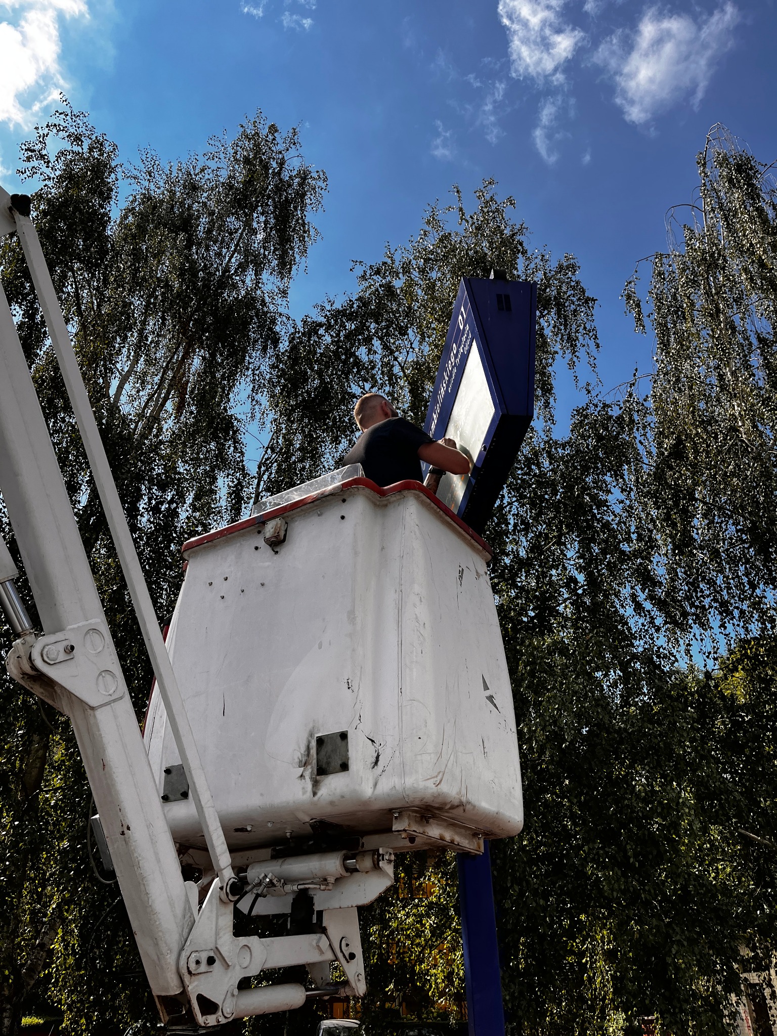 Pracownik w koszu podnośnika montuje tablicę informacyjną w Płocku, widok z dołu na tle błękitnego nieba i drzew.