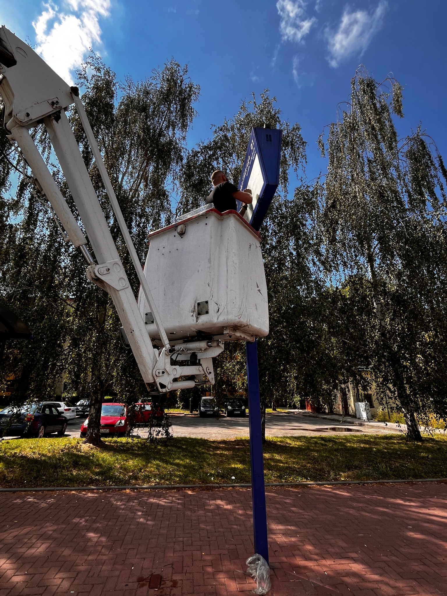 Pracownik w koszu podnośnika teleskopowego, naprawiający lub czyszczący tablicę informacyjną na tle drzew i błękitnego nieba.