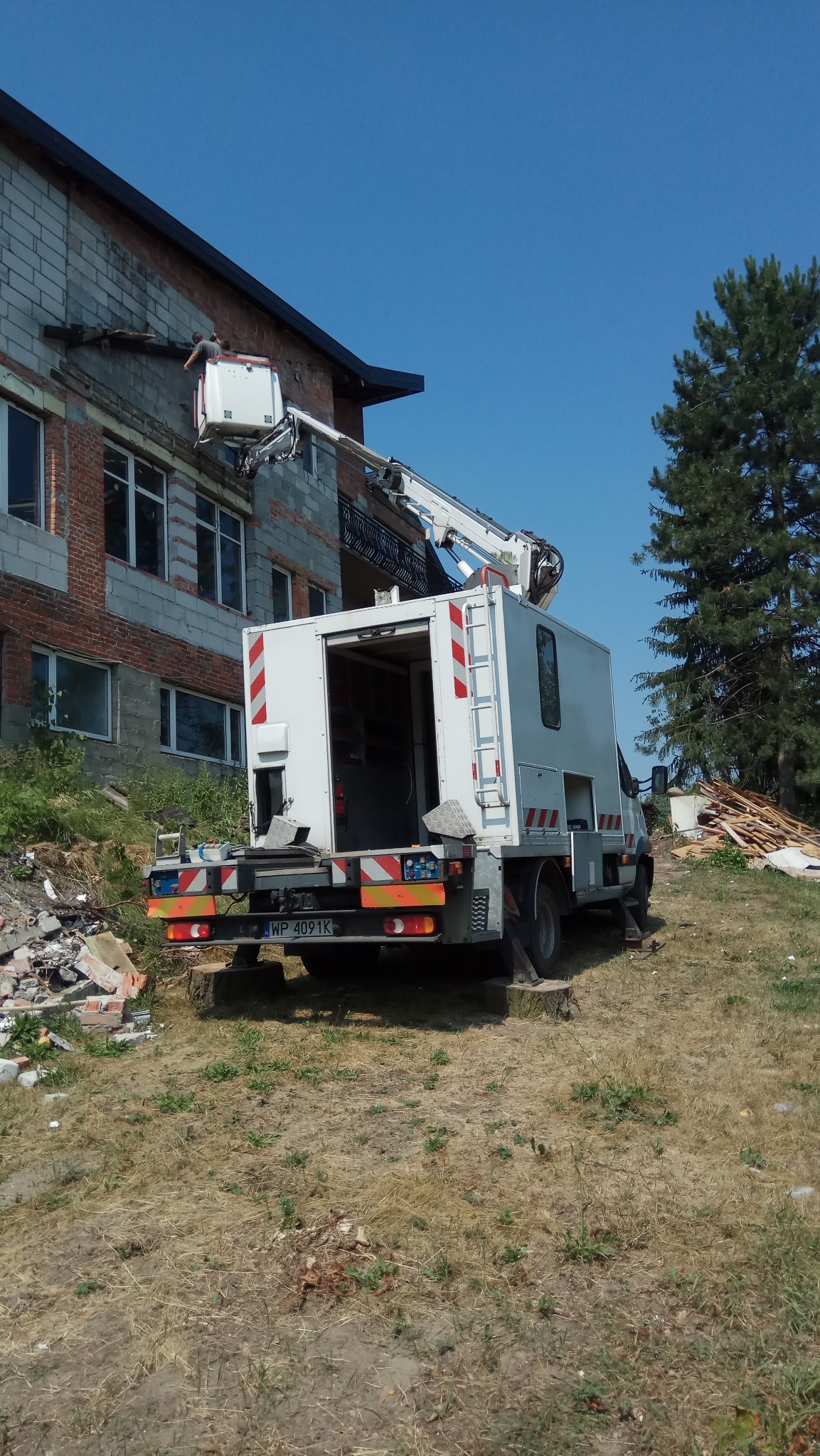 Pracownik w koszu podnośnika usuwa elementy z elewacji budynku, widoczny gruz i podnośnik koszowy z otwartym tyłem.