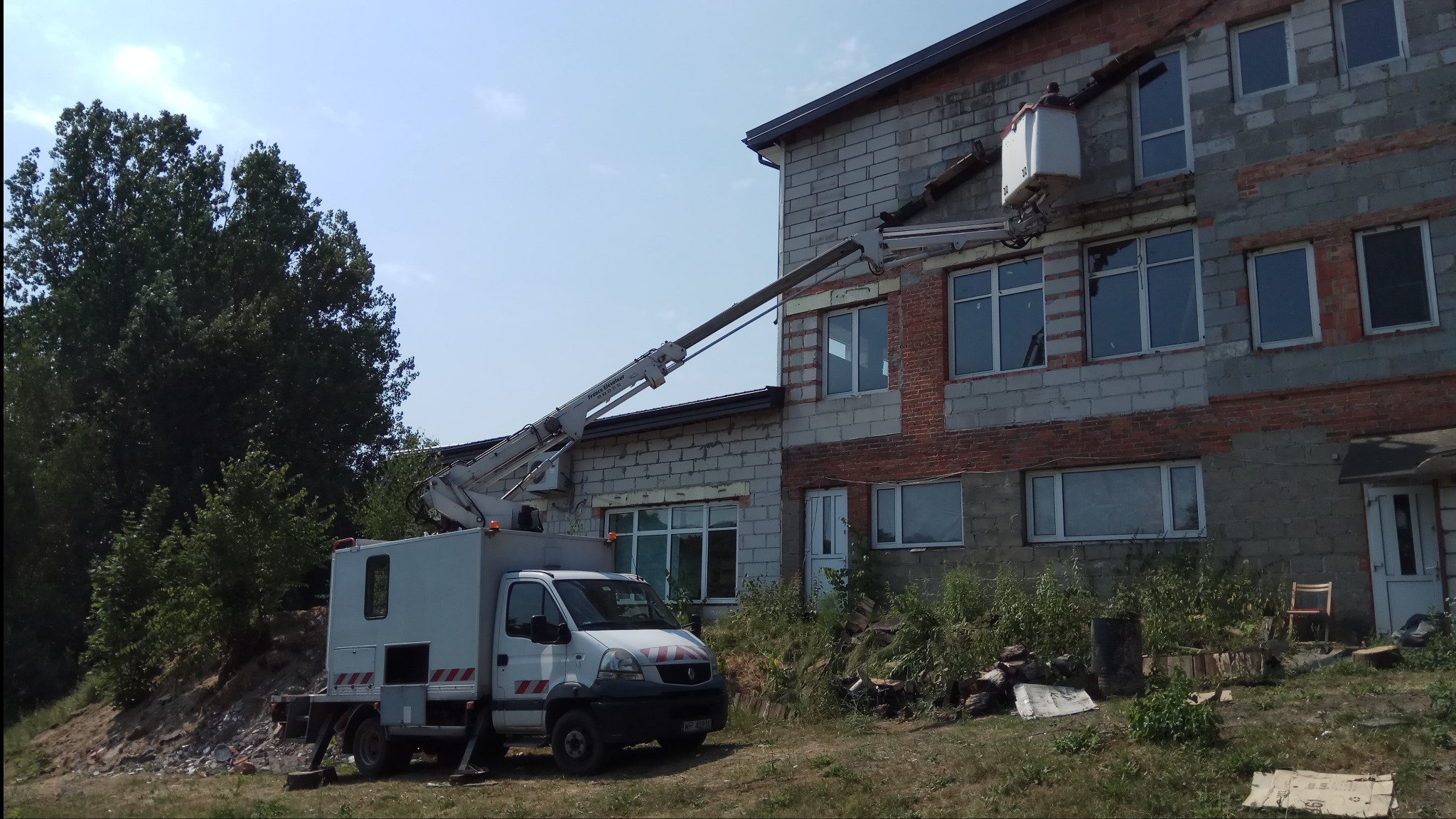 Biały podnośnik koszowy na tle dwukondygnacyjnego budynku z szarej cegły, częściowo wyremontowanego, z widoczną elewacją w trakcie prac.