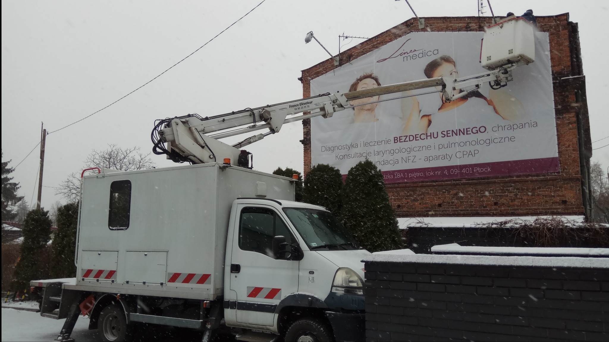 Biały podnośnik koszowy na tle ceglanego budynku z reklamą Linea Medica w zimowej scenerii, podczas opadów śniegu.