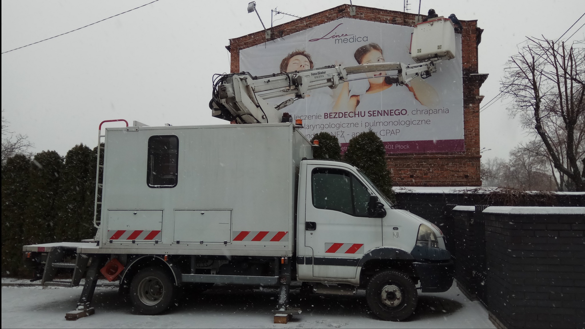 Biały samochód z podnośnikiem koszowym na tle budynku z reklamą medyczną podczas opadów śniegu. Widoczne dwie osoby w koszu podnośnika pracujące przy billboardzie.