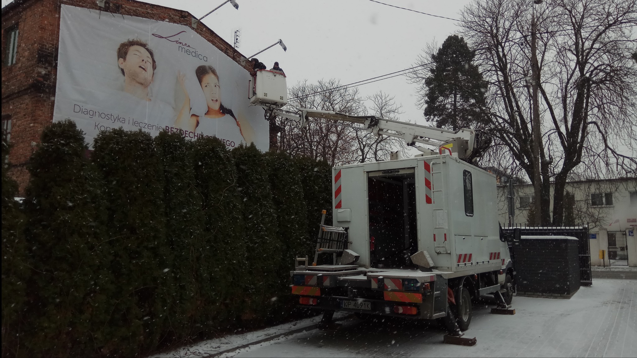 Pracownicy w koszu podnośnika montują reklamę na budynku, widoczny fragment reklamy z wizerunkami osób, śnieg pada.