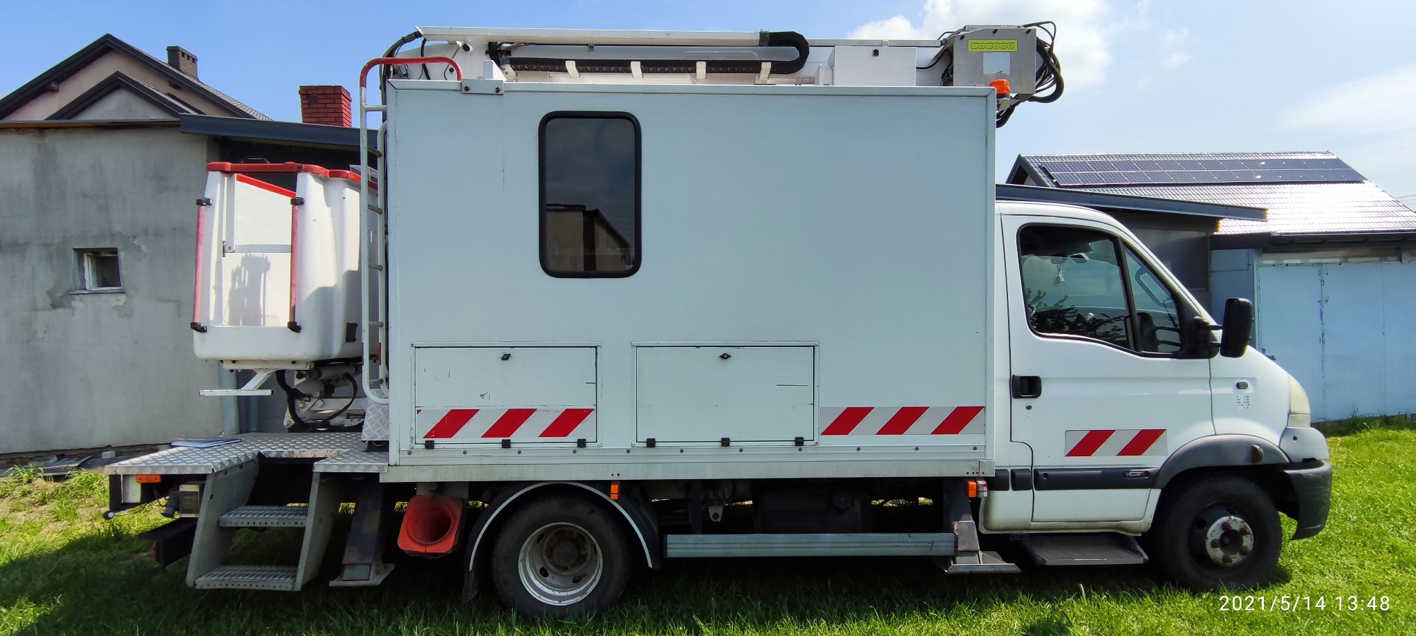 Biały samochód z podnośnikiem koszowym na tle budynków z panelami słonecznymi, widok z boku na trawniku.
