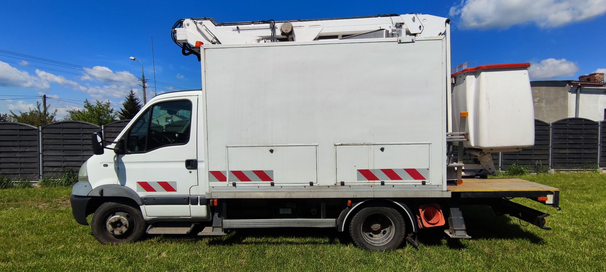 Biały samochód z podnośnikiem koszowym na tle zielonego trawnika i błękitnego nieba, widoczne czerwono-szare pasy ostrzegawcze na bokach pojazdu.