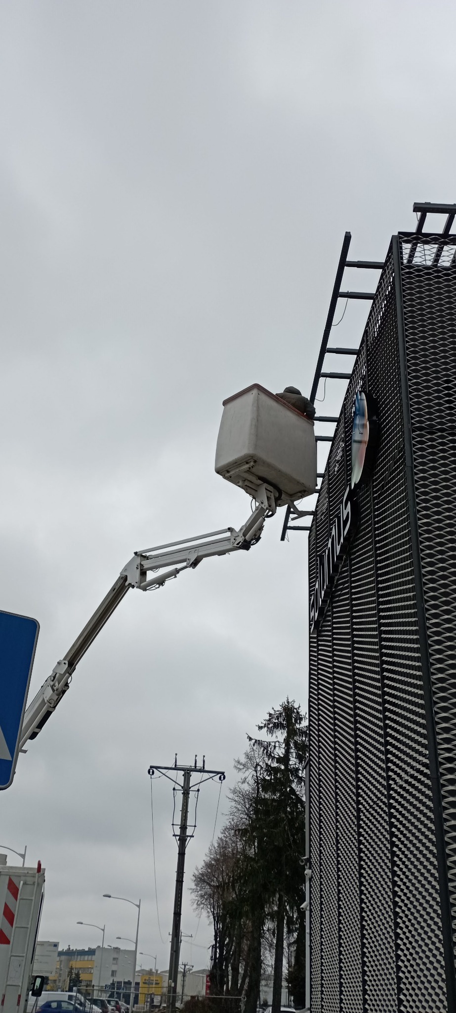 Pracownik w podnośniku koszowym na wysokości, montujący lub naprawiający element elewacji budynku z metalową siatką i napisem 'solumus'.