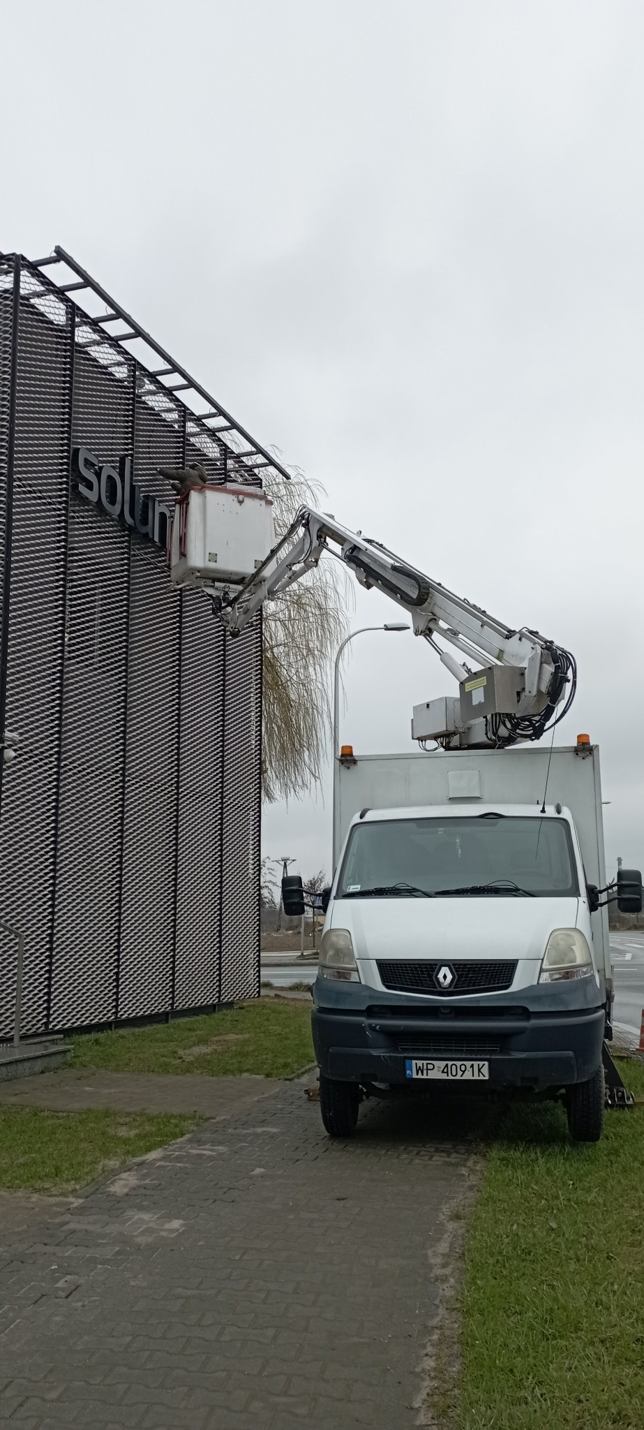 Biały samochód z podnośnikiem koszowym przy budynku z napisem 'solum'