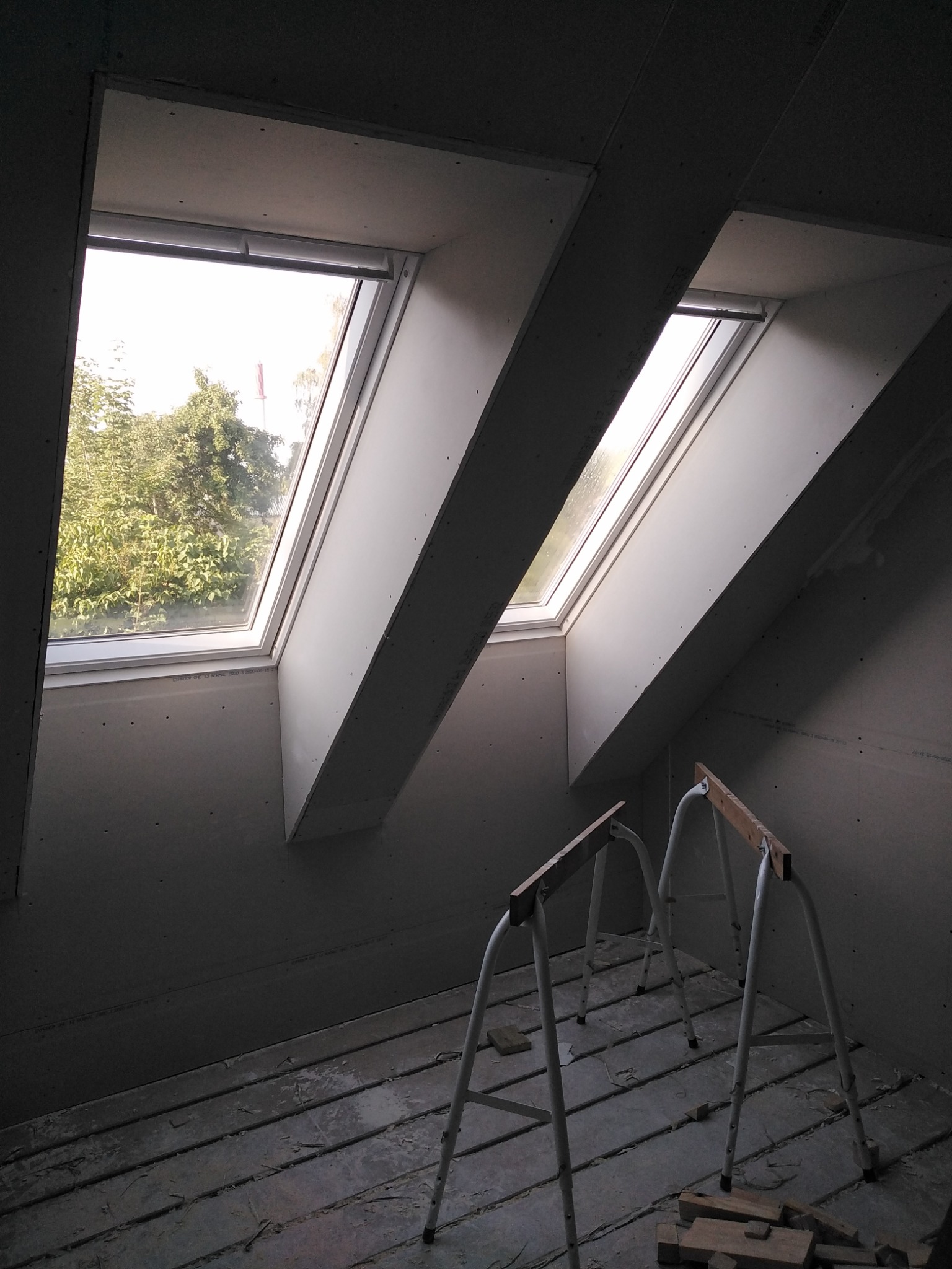 Montaż okien dachowych w poddaszu, widoczne stelaże i płyty gipsowo-kartonowe przed szpachlowaniem, drewniane deski podłogowe.