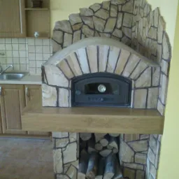 Wykończony kominek z obudową z kamienia w Dobrodzieniu, z zapasem drewna pod paleniskiem, wkomponowany w kuchenną zabudowę.