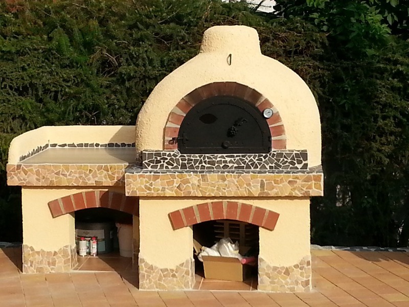 Ogród z piecem chlebowym w stylu rustykalnym, z czarnymi drzwiczkami i termometrem, wykończony mozaiką z kamienia naturalnego, na tle zielonych krzewów.