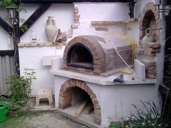 Budowa pieca chlebowego z cegły w ogrodzie, widoczne narzędzia, zaprawa murarska i fragmenty cegieł, w tle elementy dekoracyjne.