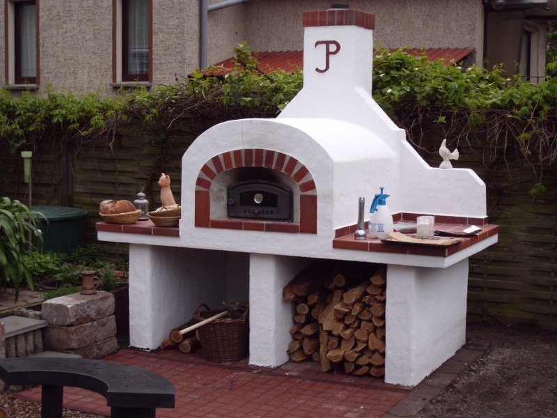 Ogród z białym, murowanym piecem chlebowym z ceglanym łukiem i kominem z inicjałami, drewnem opałowym, koszykami z chlebem i dekoracjami.