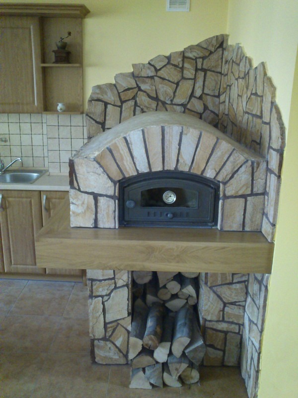 Wykończony kominek z obudową z kamienia w Dobrodzieniu, z zapasem drewna pod paleniskiem, wkomponowany w kuchenną zabudowę.