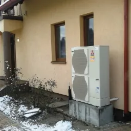 Zewnętrzna jednostka klimatyzacji, model Mitsubishi, zamontowana na betonowym fundamencie przy ścianie domu z widocznymi oknami i fragmentem rynny, na lekko ośnieżonym podłożu z kostki brukowej.