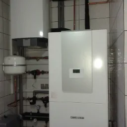 Biały piec Stiebel Eltron z podłączonymi rurami miedzianymi i szarymi, zamontowany na ścianie wyłożonej białymi płytkami.