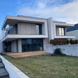 Nowoczesny dom z jasną elewacją i drewnianym tarasem na poziomie parteru, widok z perspektywy ogrodu w pochmurny dzień.