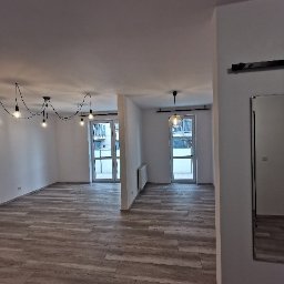 Telzarbud - Jasne, przestronne wnętrze z panelami podłogowymi imitującymi drewno, minimalistycznym oświetleniem w stylu industrialnym i widokiem na balkon przez przeszklone drzwi.