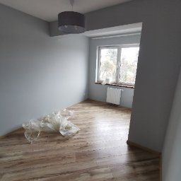 Telzarbud - Pokój po świeżym malowaniu na szaro, z ułożonymi panelami podłogowymi, zabezpieczonymi folią ochronną w rogu, widoczne okno z grzejnikiem.