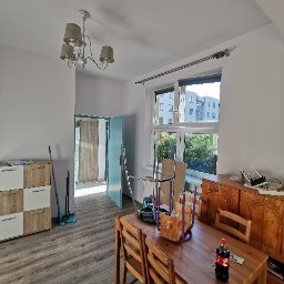 Telzarbud - Pokój przed remontem: drewniany stół z krzesłami, regał z białymi frontami, otwarte turkusowe drzwi, widoczne narzędzia i akcesoria malarskie, podłoga wyłożona panelami.