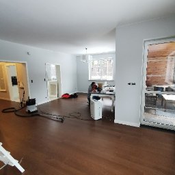 Telzarbud - Salon po ułożeniu ciemnobrązowych paneli podłogowych, widoczny pracownik sprzątający podłogę odkurzaczem przemysłowym, białe ściany i okna.