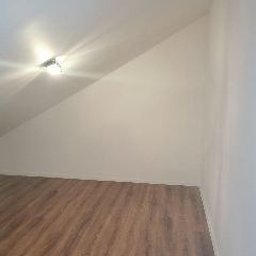 Telzarbud - Poddasze po remoncie: ściana pomalowana na biało, widoczny skos, na suficie lampa halogenowa, na podłodze panele w kolorze jasnego drewna z wyraźnym rysunkiem słojów.