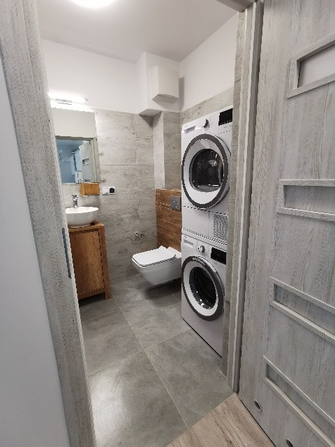 Nowoczesna łazienka z szarymi płytkami, wiszącą toaletą, umywalką na drewnianej szafce i pralko-suszarką ustawioną jedna na drugiej, widok zza uchylonych drzwi.